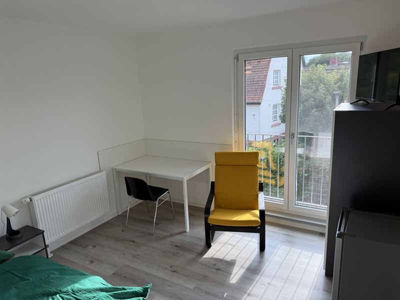 Helles Zimmer mit Schreibtisch, Sessel und bodentiefem Fenster