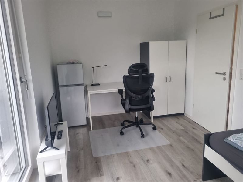 Zimmer mit Schreibtisch, ergonomischem Bürostuhl, Fernseher, Kühlschrank und Kleiderschrank